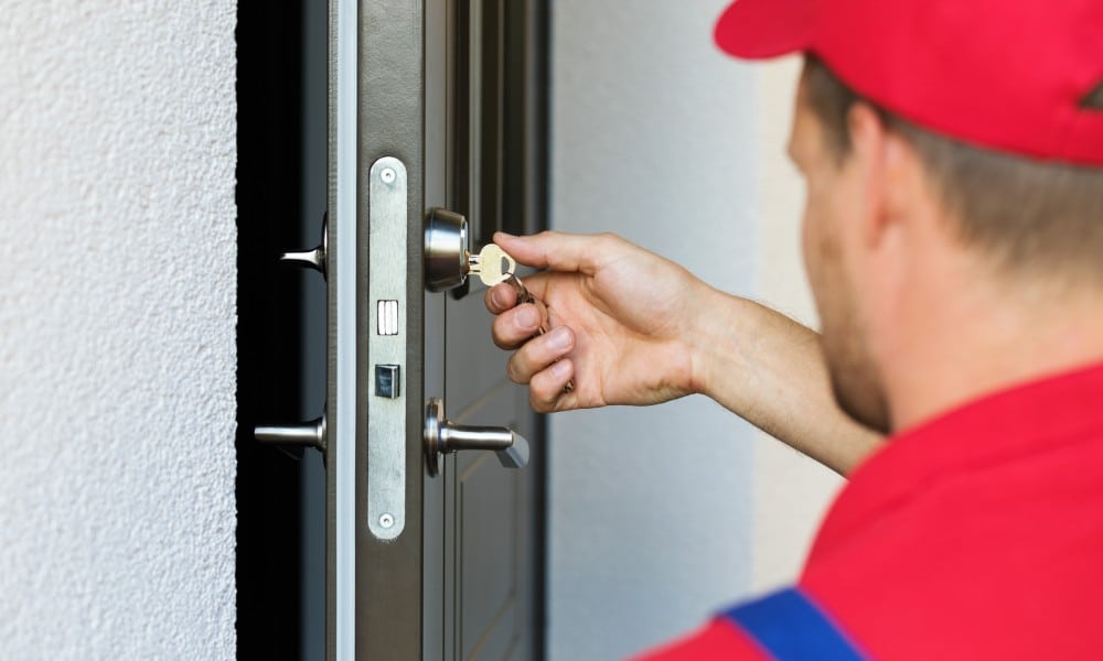 A man in a red hat and shirt and blue overalls holds a key on a keyring in the lock of a slightly open door.